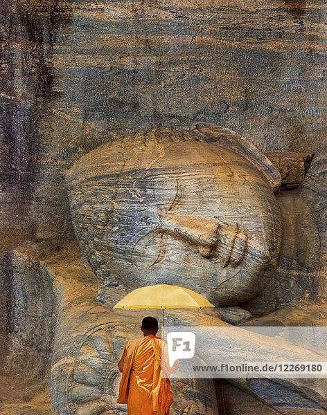 Rückansicht eines Mannes in orangefarbener Robe,  der vor einer riesigen,  liegenden,  in den Fels gehauenen Buddha-Skulptur steht.