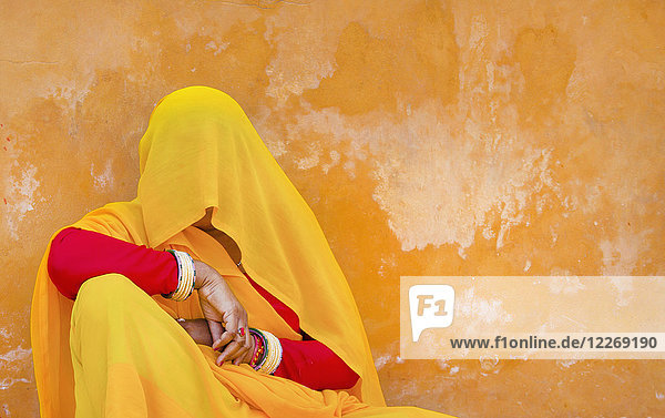 Frau mit rot-gelbem Sari und Schleier  der ihren Kopf bedeckt  auf dem Boden sitzend  an orangefarbene Wand gelehnt.