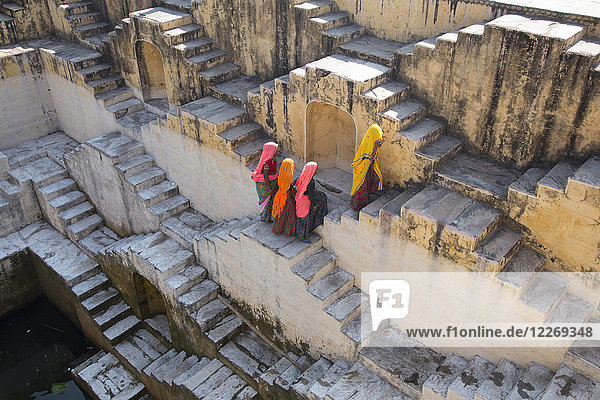 Hochwinkelaufnahme von vier Saris tragenden Frauen  die die Treppe um den Wasserpool eines alten Gebäudes hinaufgehen.