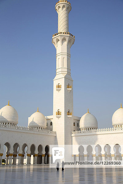 Rückansicht zweier vor der Moschee stehender Frauen mit weiß getünchter Kolonnade  Minarett und Kuppeln.