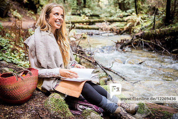 Young woman relaxing on forest riverbank with notebook