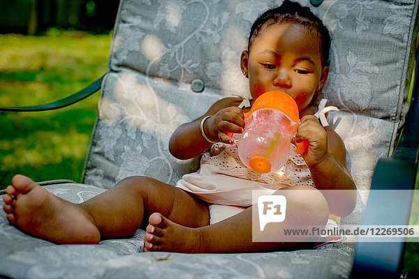 Baby girl sitting on garden lounge chair drinking from baby cup
