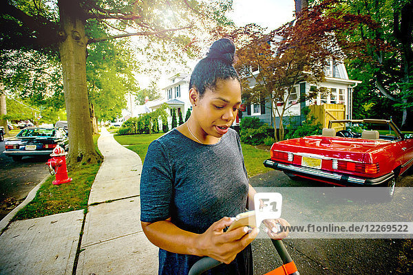 Mid adult woman with pushchair on suburban sidewalk looking at smartphone