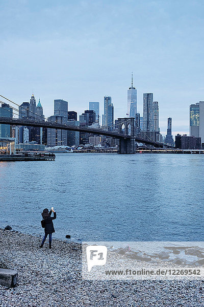 Frau fotografiert die Brooklyn Bridge und die Skyline von Lower Manhattan vom Flussufer aus  New York  USA
