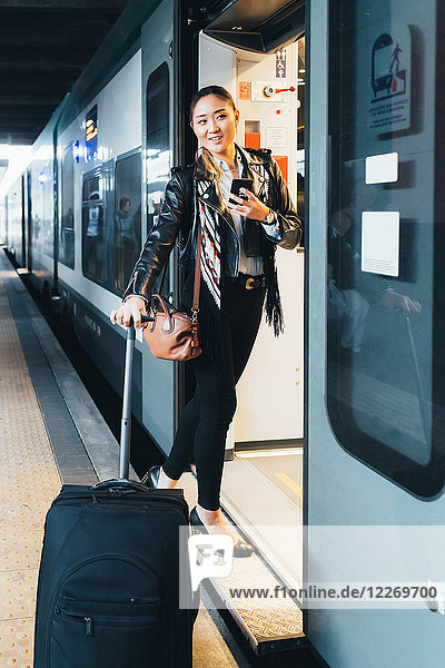 Frau steigt in den Zug ein  zieht einen Koffer mit Rädern  hält ein Smartphone