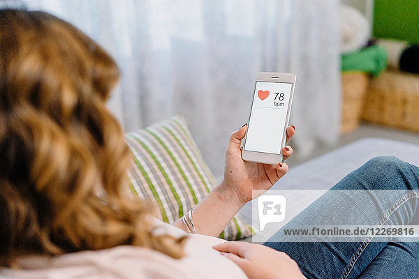 Schwangere Frau auf dem Sofa mit Blick auf die Herzfrequenz auf dem Smartphone  Blick über die Schulter