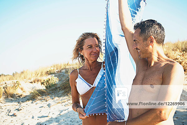 Erwachsenes Paar am Strand  hält Stranddecke zwischen sich und lächelt