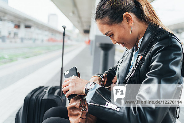 Frau sitzt auf Bahnsteig  schaut durch Handtasche  Koffer neben ihr  hält Smartphone