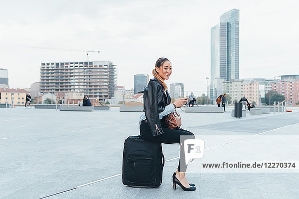 Porträt einer Geschäftsfrau im Freien  die auf einem Koffer sitzt und lächelt