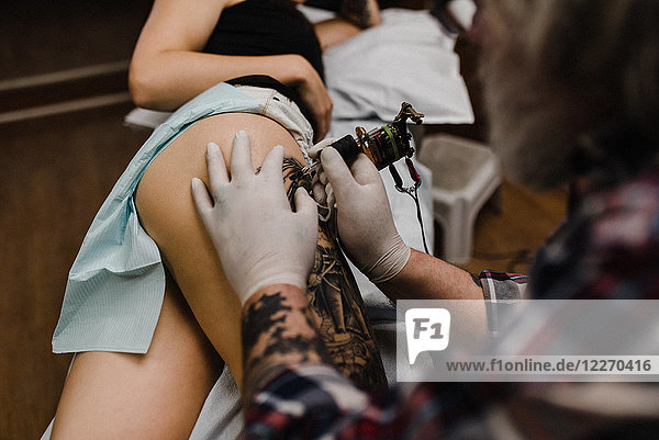 Tattooist tattooing young woman's thigh  close-up