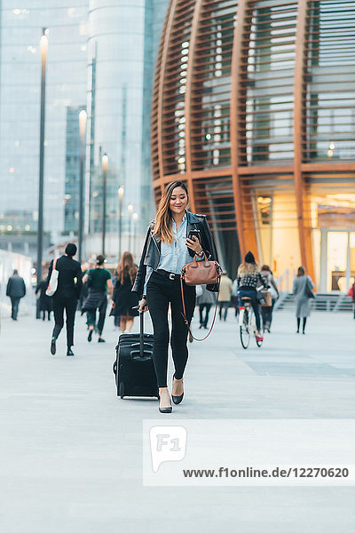 Geschäftsfrau geht im Freien spazieren  zieht Rollkoffer  hält Smartphone und Handtasche und lächelt