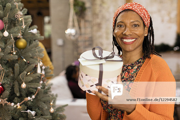 Portrait lächelnde  selbstbewusste Frau mit Geschenk neben dem Weihnachtsbaum