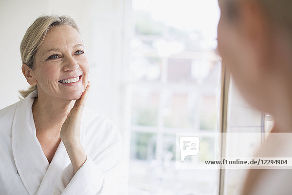 Lächelnde reife Frau berührt Gesicht im Badezimmerspiegel