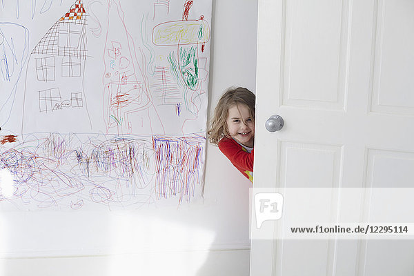 Porträt eines fröhlichen Mädchens  das sich hinter der Tür versteckt  gegen das Zeichnen an der Wand.