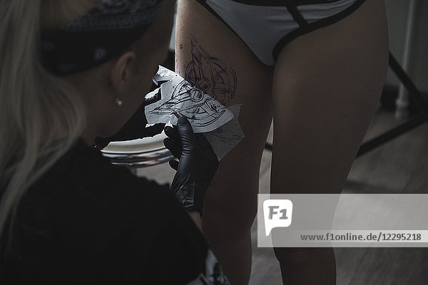 Künstlerin beim Abziehen der Tattoo-Schablone am Oberschenkel der Kundin im Atelier