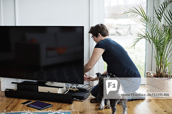 Pregnant woman arranging cables of television set while kneeling by dog in living room