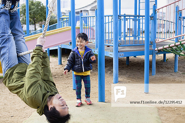 Lächelnder Junge schaut Vater an  wie er auf dem Spielplatz am Seil schwingt.