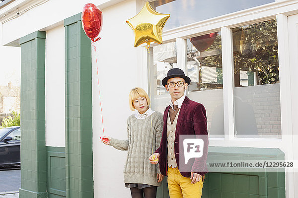 Young couple holding balloons and looking away while standing against window