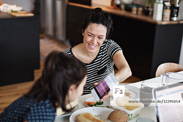 Smiling mother showing mobile phone to daughter while having breakfast at dining table