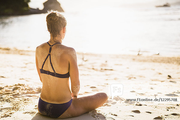 Rückansicht der mittleren erwachsenen Frau im Bikini beim Meditieren am Strand an einem sonnigen Tag.