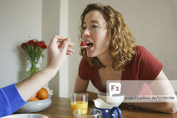 Hand of man feeding strawberry to woman at table in room