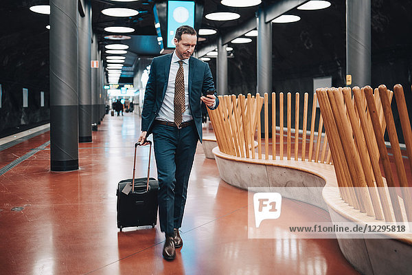 Full length of businessman with luggage and mobile phone walking in railroad platform