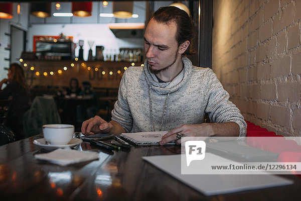 Junger Mann beim Sitzen mit Handy und Buch auf dem Tisch im Cafe