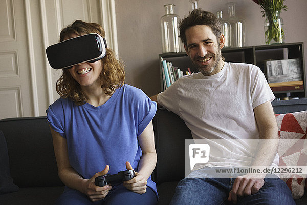 Porträt eines lächelnden Mannes,  der von einer Frau auf einem Virtual-Reality-Headset im Wohnzimmer sitzt.