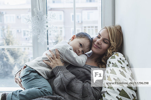 Smiling young mother holding son by window sill at home