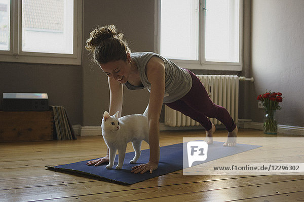 Lächelnde Frau übt Plankenposition über weißer Katze auf Trainingsmatte zu Hause