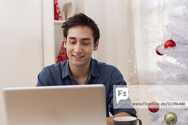 Lächelnder junger Mann mit Laptop beim Sitzen am künstlichen Weihnachtsbaum zu Hause