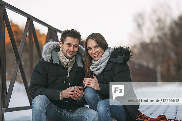 Porträt eines glücklichen Paares  das im Winter mit Getränken an der Strandpromenade sitzt.