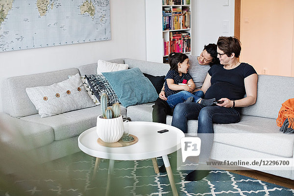Smiling lesbian couple sitting with daughter on sofa in living room at home