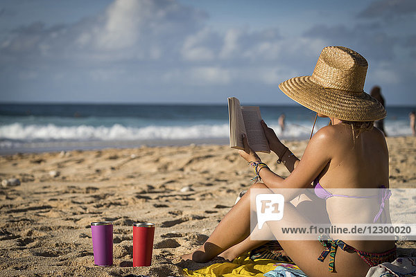 Frau in Bikini und Sonnenhut sitzt und liest ein Buch am Strand  Oahu  Hawaii-Inseln  USA