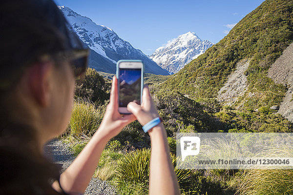 Blick über die Schulter einer jungen Frau  die den Mount Cook mit ihrem Smartphone fotografiert  Hooker Valley Track  Canterbury  Neuseeland