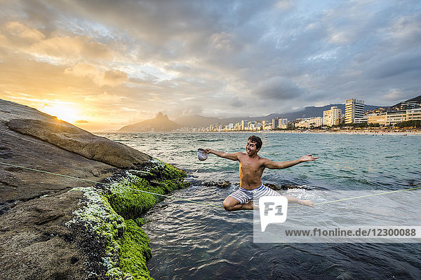 Young man looking at camera while balancing with smile on slackline tensioned over coastal water at sunset  Rio de Janeiro  Brazil