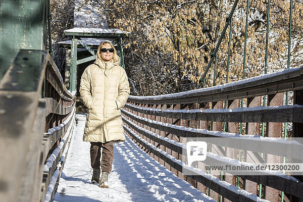 Erwachsene Frau mit Sonnenbrille  die mit den Händen in den Taschen über eine schneebedeckte Flussbrücke läuft  Durango  Colorado  USA