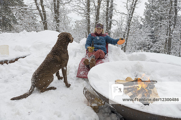 Woman relaxing on snow by burning fire pit and playing with dog  Durango  Colorado  USA