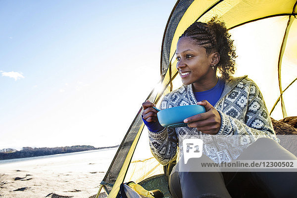 Young woman sitting in tent and eating breakfast while camping on coastal beach  Newburyport  Massachusetts  USA