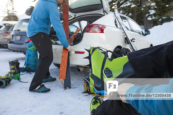 Two backcountry skiers preparing for skiing with one putting on ski boots and the other near car trunk