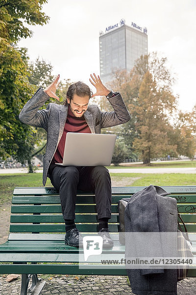 Hispanischer Mann  der eine Errungenschaft mit Laptop auf einer Bank in einem öffentlichen Park mit einem Geschäftsgebäude im Hintergrund feiert