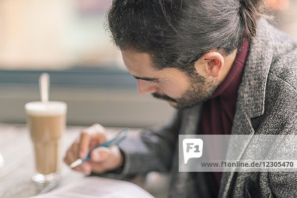 Ein hispanischer Mann  der in einem Café sitzt und schreibt  trägt einen eleganten Freizeitblazer und einen Rollkragenpullover