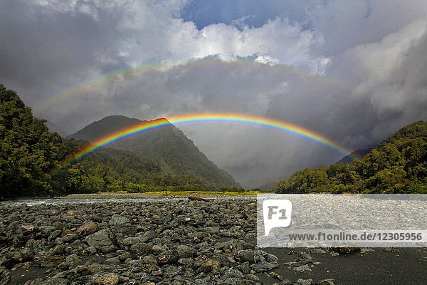 Doppelter Regenbogen vor großen Wolken über felsigem Tal  Neuseeland