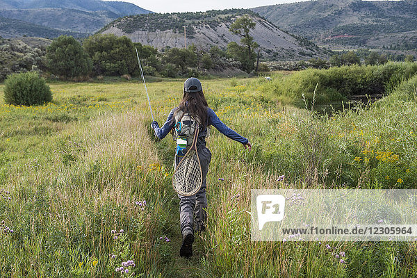 Female angler walking through meadow with fishing rod in hand  Colorado  USA