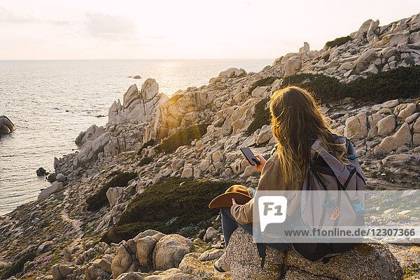 Italien  Sardinien  Frau auf einer Wanderung auf einem Felsen an der Küste sitzend mit Handy