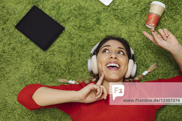 Fröhliche junge Frau liegt auf dem Teppich und hört Musik mit Kopfhörern.