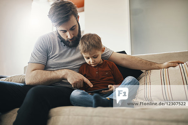 Father and little son sitting together on the couch looking at smartphone