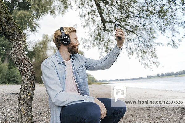 Deutschland Düsseldorf Mann mit Kopfhörer nimmt Selfie am Strand mit Smartphone
