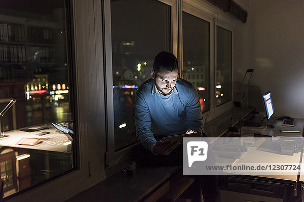Businessman sitting on window sill in office at night using tablet