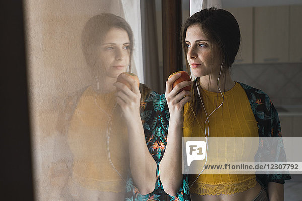 Junge Frau mit Kopfhörer und Apfel aus dem Fenster schauend